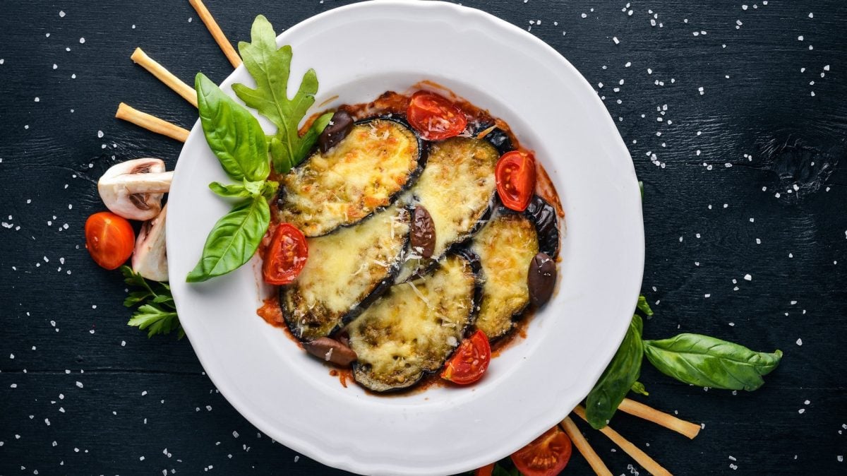 Gebackene Aubergine mit Tomaten auf weißem Teller, angerichtet auf dunklem Untergrund