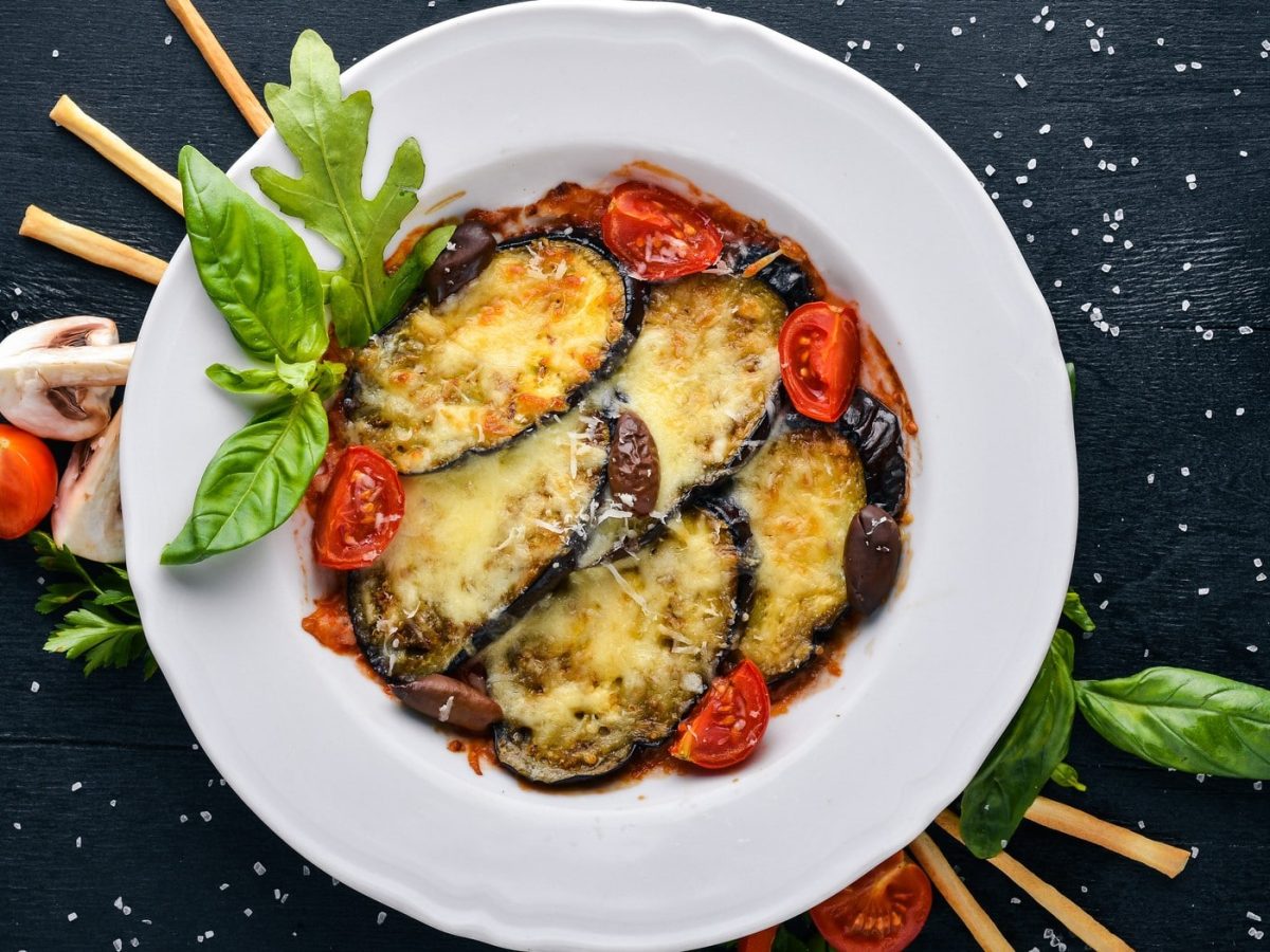 Gebackene Aubergine mit Tomaten auf weißem Teller, angerichtet auf dunklem Untergrund