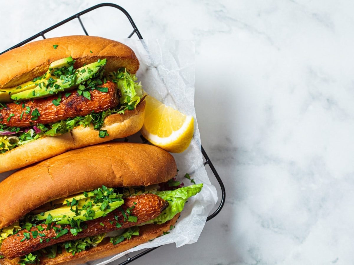 Vegane Hotdogs auf Backpapier auf einem schwarzen Gitter. Daneben liegt eine Zitrone.