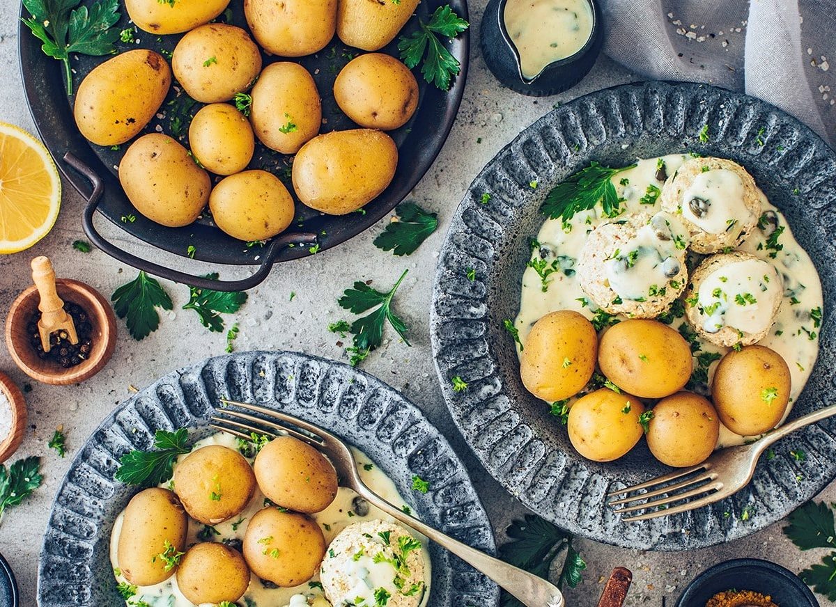 Zwei Teller vegane Königsberger Klopse mit Pellkartoffeln in der Draufsicht, außerdem ein Topf Kartoffeln und Schalen mit Gewürzen.