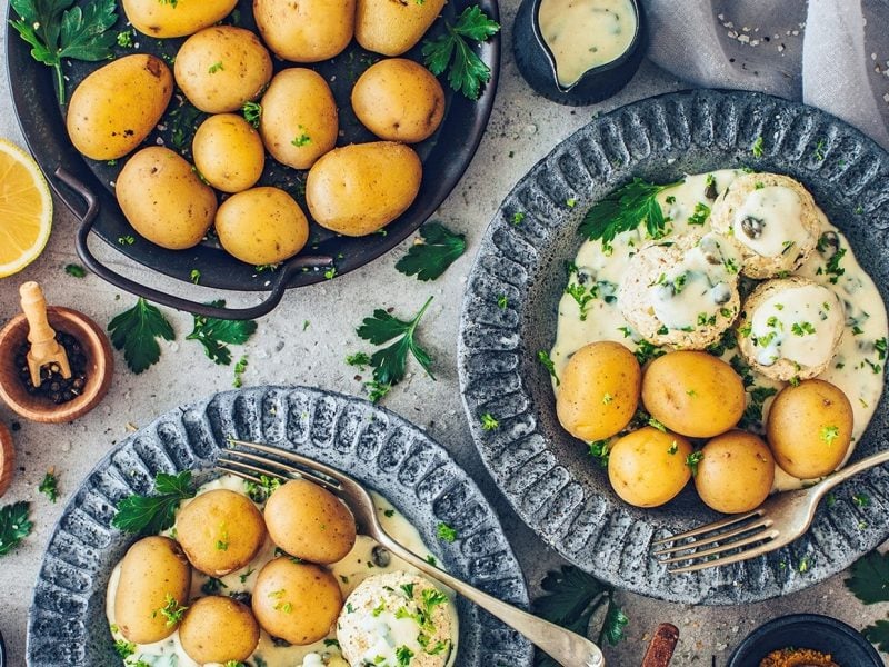 Zwei Teller vegane Königsberger Klopse mit Pellkartoffeln in der Draufsicht, außerdem ein Topf Kartoffeln und Schalen mit Gewürzen.