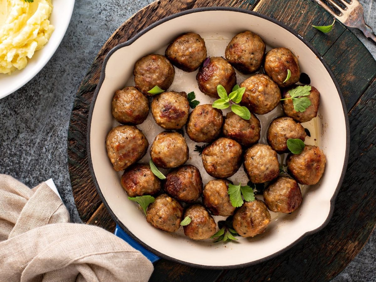 Eine Pfanne vegane Köttbullar mit Kräutern bestreut auf einem runden Brett in der Draufsicht, daneben eine Schale Kartoffelpüree, ein Geschirrtuch und eine Gabel.