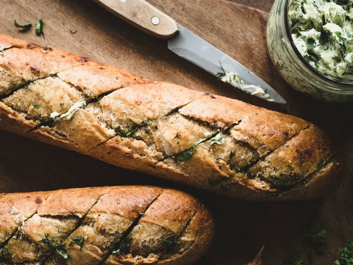 zwei vegane Kräuterbaguettes in der Draufsicht, daneben ein Messer und ein Glas Kräuterbutter.