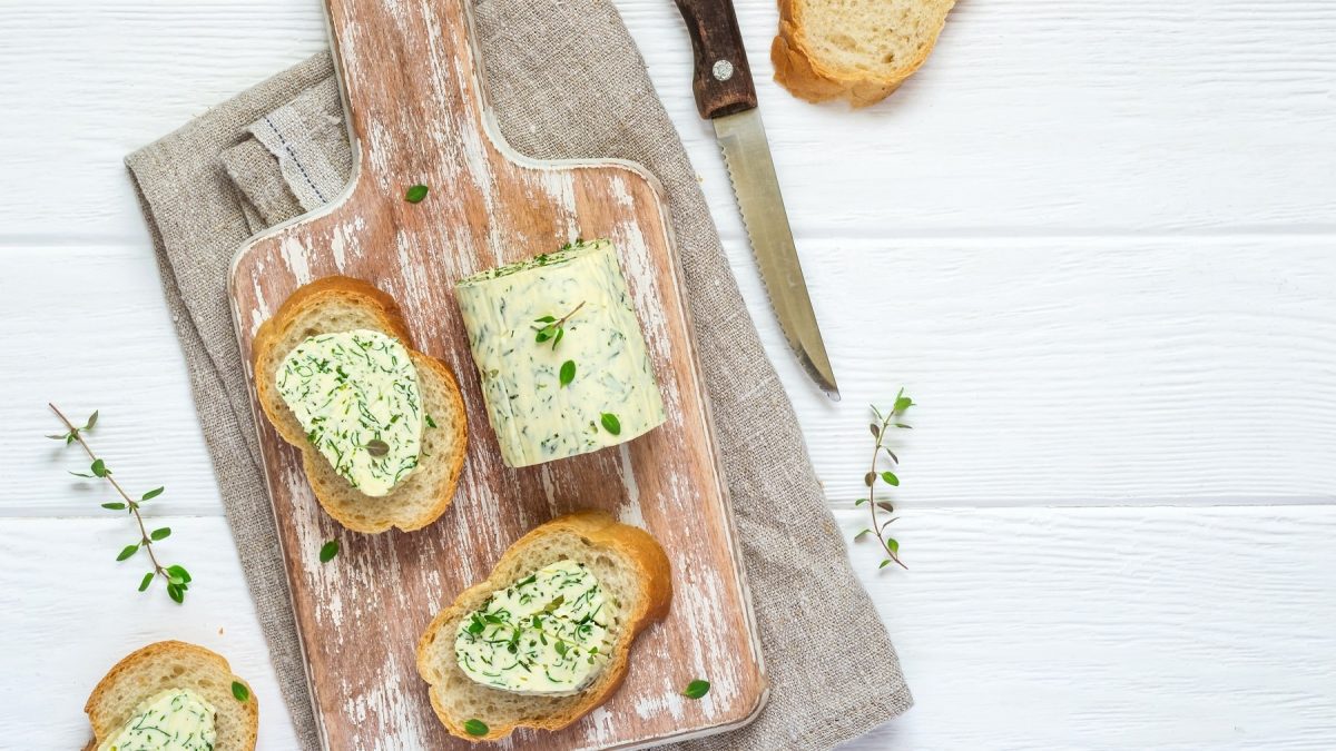 Brotscheiben mit veganer Kräuterbutter auf einem Brett in der Draufsicht, daneben liegen eine Rolle Kräuterbutter, frische Kräuter und ein Messer.