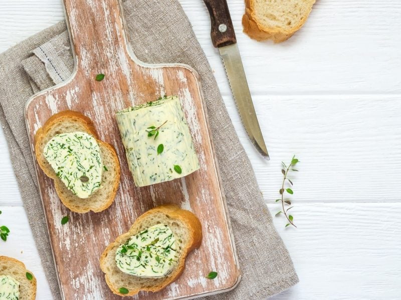 Brotscheiben mit veganer Kräuterbutter auf einem Brett in der Draufsicht, daneben liegen eine Rolle Kräuterbutter, frische Kräuter und ein Messer.