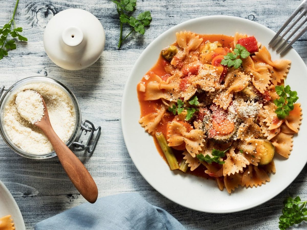 Ein Teller vegane Pasta mit Gemüseragout in der Draufsicht, daneben eine Gabel und ein Glas mit Parmesan, außerdem frische Kräuter und ein Salzstreuer.