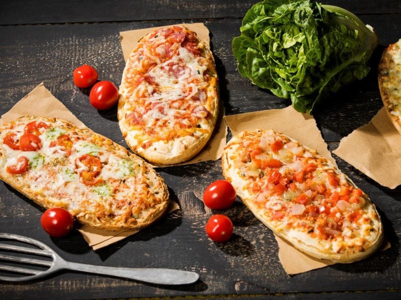 Drei vegane Pizzabrötchen auf Backpapier, daneben liegen kleine Tomaten und ein Pfannenwender.