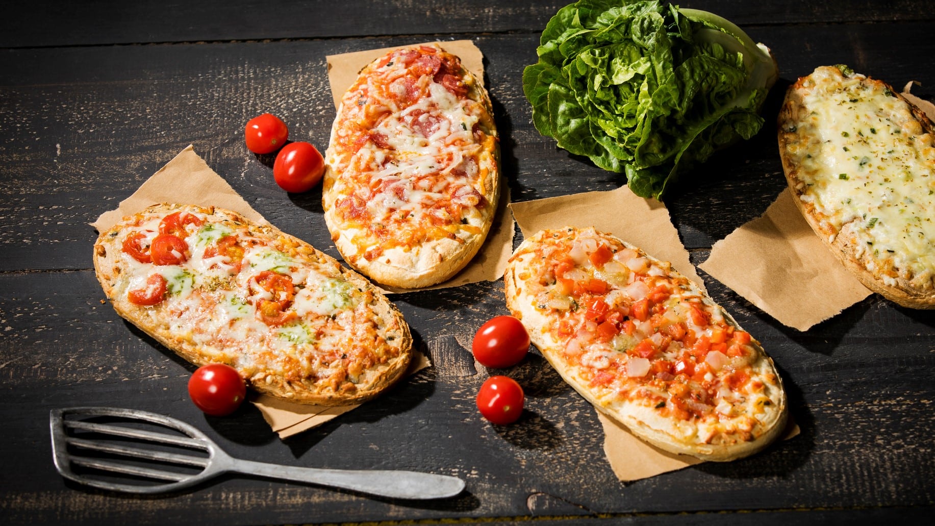 Drei vegane Pizzabrötchen auf Backpapier, daneben liegen kleine Tomaten und ein Pfannenwender.