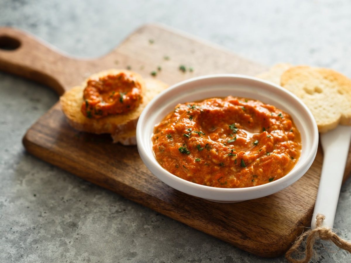 Eine Schale veganer Tomate-Walnuss-Aufstrich steht auf einem Brett, auf dem auch zwei bestrichene Baguettescheiben liegen.