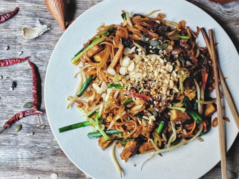 Ein Teller veganes Pad Thai mit Erdnüssen, Frühlingszwiebeln und Möhren, daneben Essstäbchen. Alles in der Draufsicht.