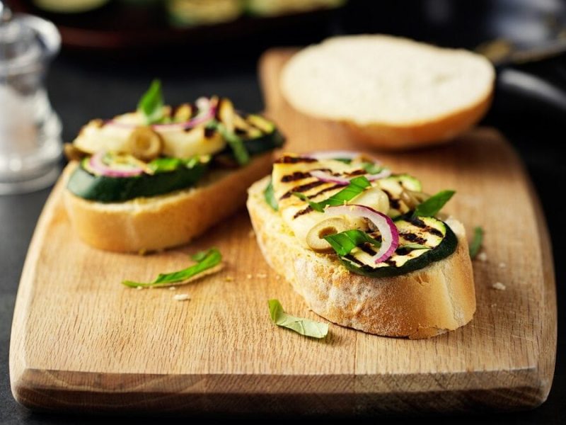 Auf einem Holzbrett liegen zwei Bruschetta sind eine klassische italienische Vorspeise. Scheiben Weißbrot werden gegrillt und darauf kommt, was schmeckt oder gerade da. Eine Kombi aus beidem ist perfekt. Klassische Beläge sind feine Tomatenstückchen, Oliven und Basilikum-Blätter. Zumindest die Oliven und das Basilikum habe es auf unsere Zucchini-Bruschetta mit Halloumi geschafft. Der Halloumi ist ein halbfester Käse aus Griechenland, der auch gern als Grillkäse bezeichnet wird. Kommt er auf den Grill, kann er wunderbar mariniert werden. Mein Rezept für die Zucchini-Bruschetta mit Halloumi ist für eine Grillpfanne gemacht. Du kannst es aber ebenso alles auf dem Grill packen, was ich in die Pfanne werfe. Interessierst du dich generell für Zutaten, deren Eigenschaften sowie Zubereitung? Schau doch mal in unserer Kochschule vorbei. Dort verraten wir zum Beispiel. ob Oliven eigentlich Obst oder Gemüse sind. Nun wünsche ich dir viel Freude bei der Zubereitung der Zucchini-Bruschetta mit Halloumi. Daneben noch eine Scheibe Weißbrot ohne Belag. Neben dem Holzbrett steht ein Salzstreuer.