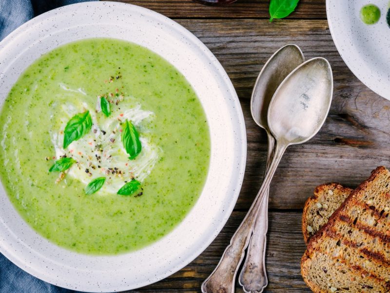 Zucchinicreme-Suppe mit Gorgonzola auf einem weißen Teller. Daneben zwei Scheiben Brot und zwei Löffel.