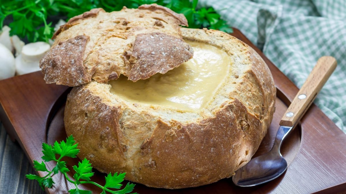 Ein Brotlaib gefüllt mit der Zwiebel-Käsesuppe auf einem dunklen Brett mit Kräutern und einem blau-weiß kariertem Tuch im Hintergrund.