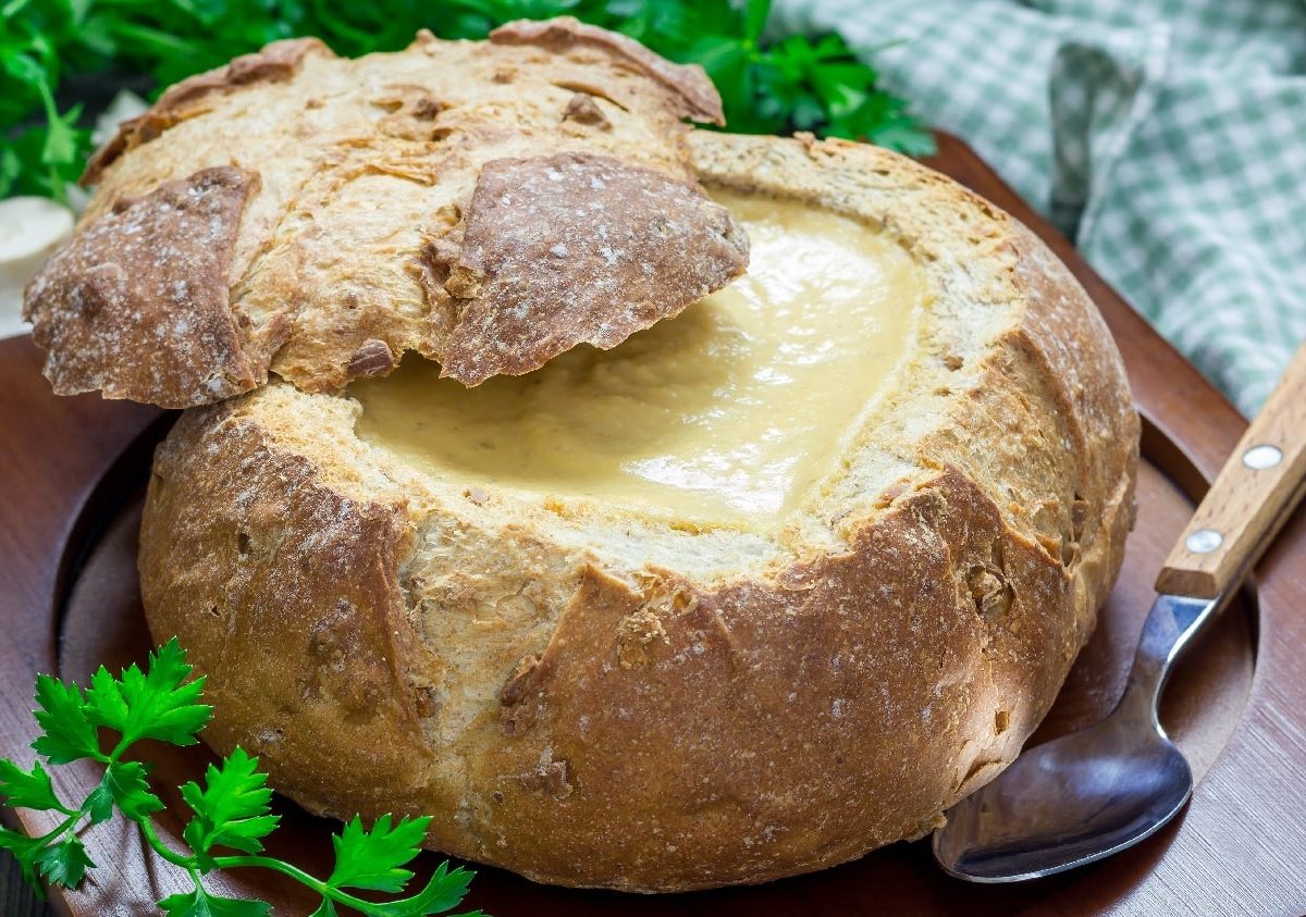 Ein Brotlaib gefüllt mit der Zwiebel-Käsesuppe auf einem dunklen Brett mit Kräutern und einem blau-weiß kariertem Tuch im Hintergrund.