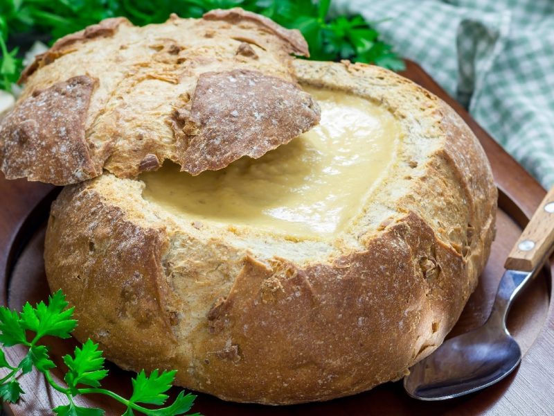 Ein Brotlaib gefüllt mit der Zwiebel-Käsesuppe auf einem dunklen Brett mit Kräutern und einem blau-weiß kariertem Tuch im Hintergrund.