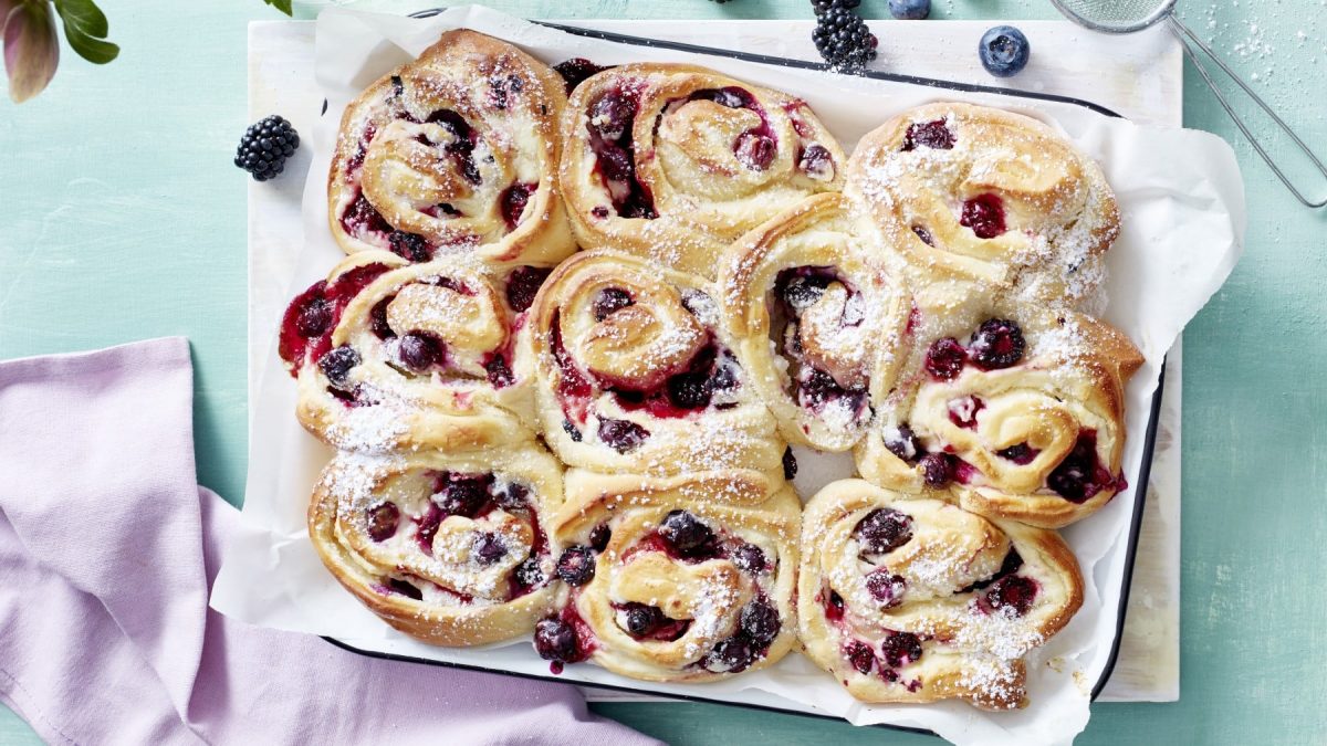 Auf einem Blech liegt ein Beeren-Schneckenkuchen. Er ist mit Puderzucker bestreut.