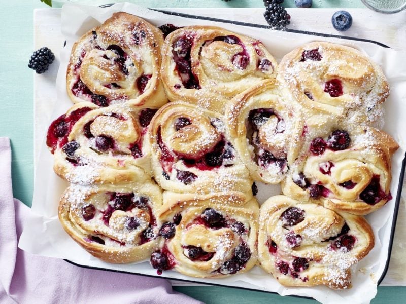 Auf einem Blech liegt ein Beeren-Schneckenkuchen. Er ist mit Puderzucker bestreut.