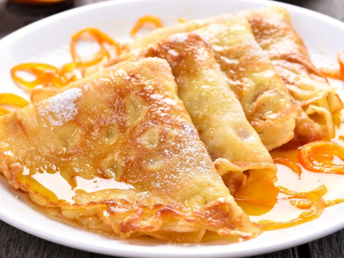 Mehrere Crêpes Suzette mit Orangenzeste auf weißem Teller.