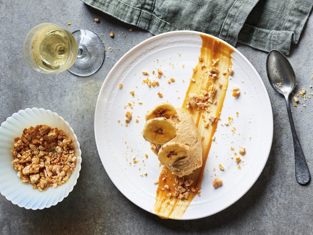 Erdnussbutterparfait mit Salted-Caramel-Crunch, serviert auf einem weißen Teller mit Bananen garniert und mit Wein auf einem Steintisch serviert.