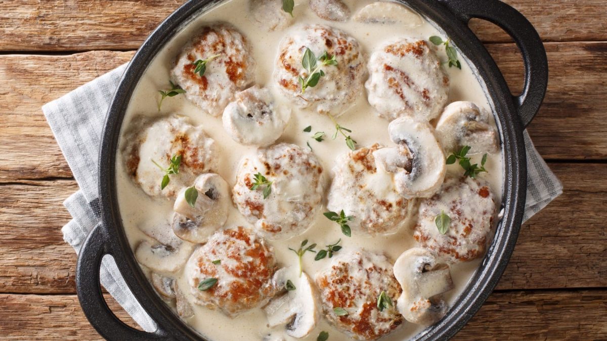 Hackbällchen in Champignonrahmsauce in schwarzer Pfanne auf hölzernem Untergrund.