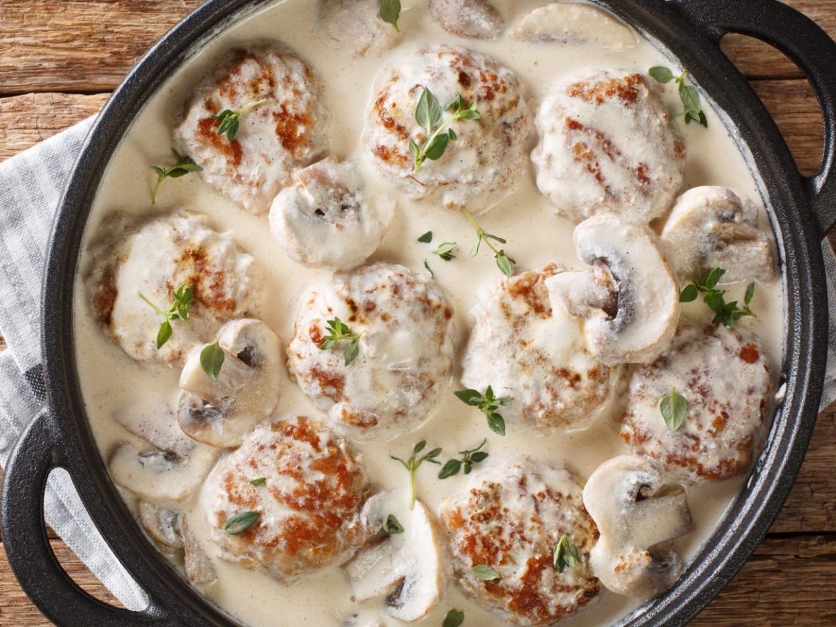 Hackbällchen in Champignonrahmsauce in schwarzer Pfanne auf hölzernem Untergrund.