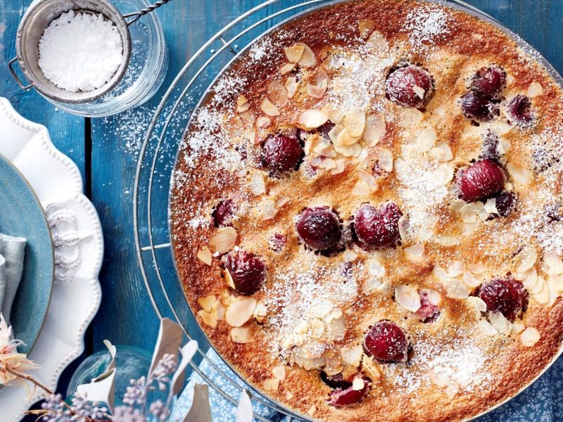 Kirsch-Clafoutis, bestehend aus einem Pfannkuchenteig und frischen Kirschen, gegossen in eine Kuchenform und ausgebacken, und bestreut mit Mandeln und Puderzucker, serviert auf einem blauen Tisch.