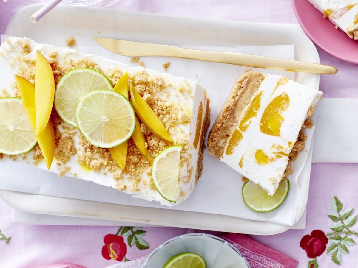 Auf einem Teller steht ein Mango-Kokos-Cheesecake. Er ist mit Limetten und Mango belegt und mit Streuseln bestreut.