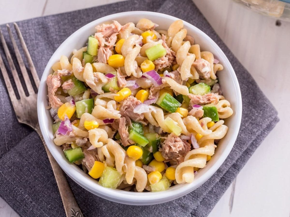 Nudelsalat mit Thunfisch, Gurke, Mais und Zwiebel in weißer Schale auf grauem Tuch.