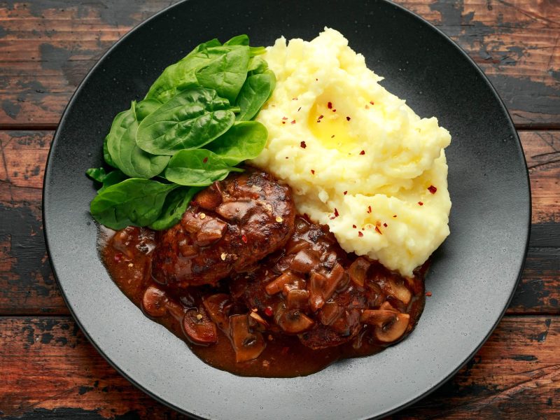 Salisbury Steak mit Kartoffelbrei auf schwarzem Teller. Dieser steht auf Holztisch.