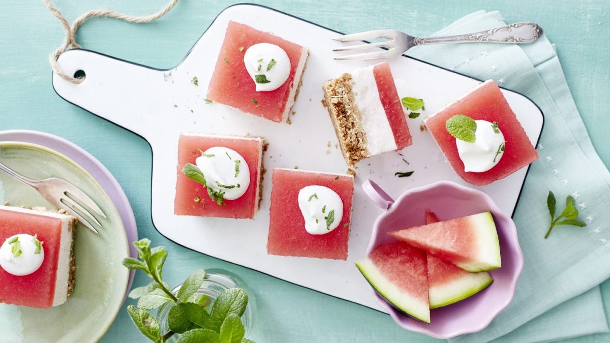 Auf einem Schneidebrett liegen mehrere Stück Wassermelonen-Kokos-Schnitten. Sie sind mit einem Klecks Schlagsahne bedeckt.