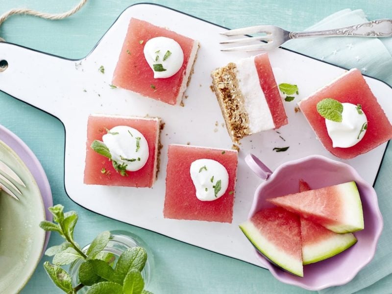 Auf einem Schneidebrett liegen mehrere Stück Wassermelonen-Kokos-Schnitten. Sie sind mit einem Klecks Schlagsahne bedeckt.