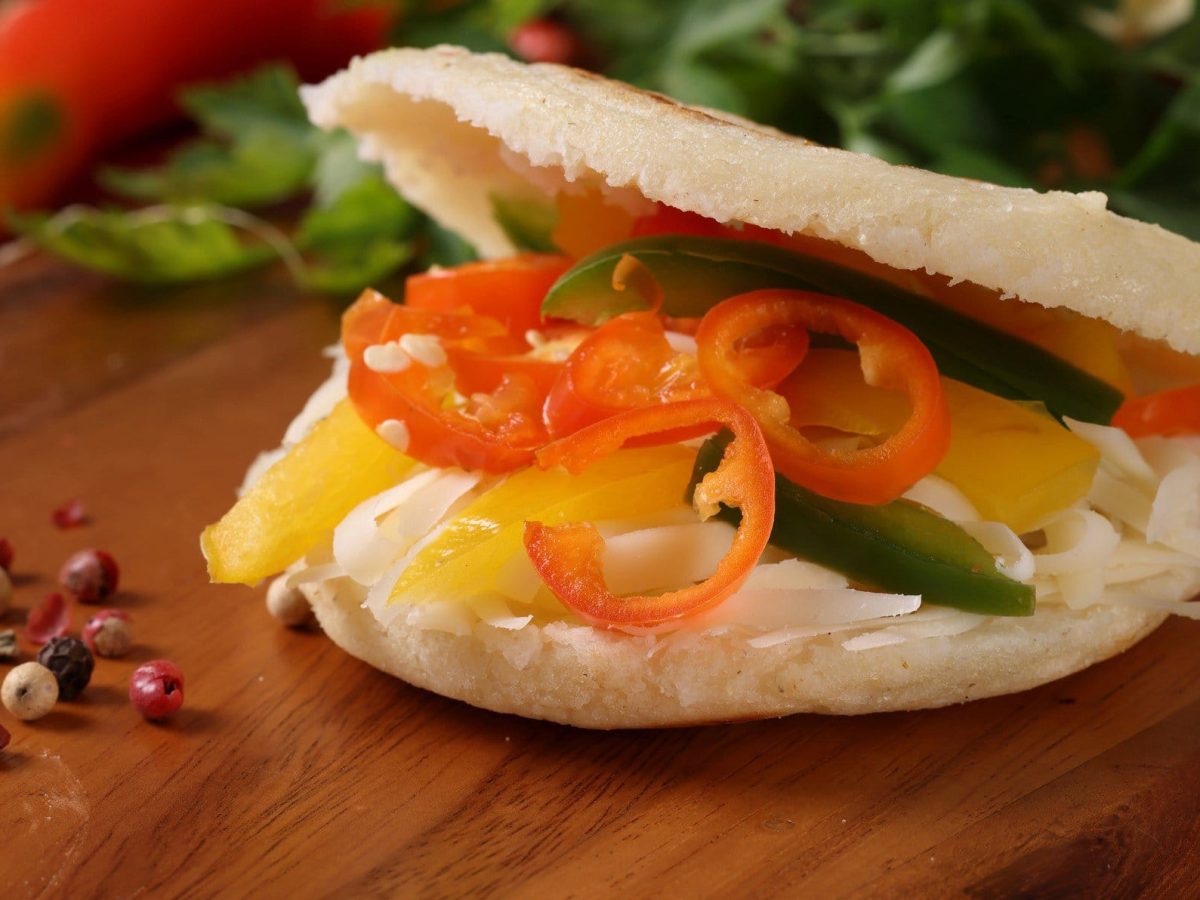 Arepas mit Chili-Kraut und unten Paprika-Streifen auf Holztisch. Daneben bunter Pfeffer, Chilis und Petersilie.