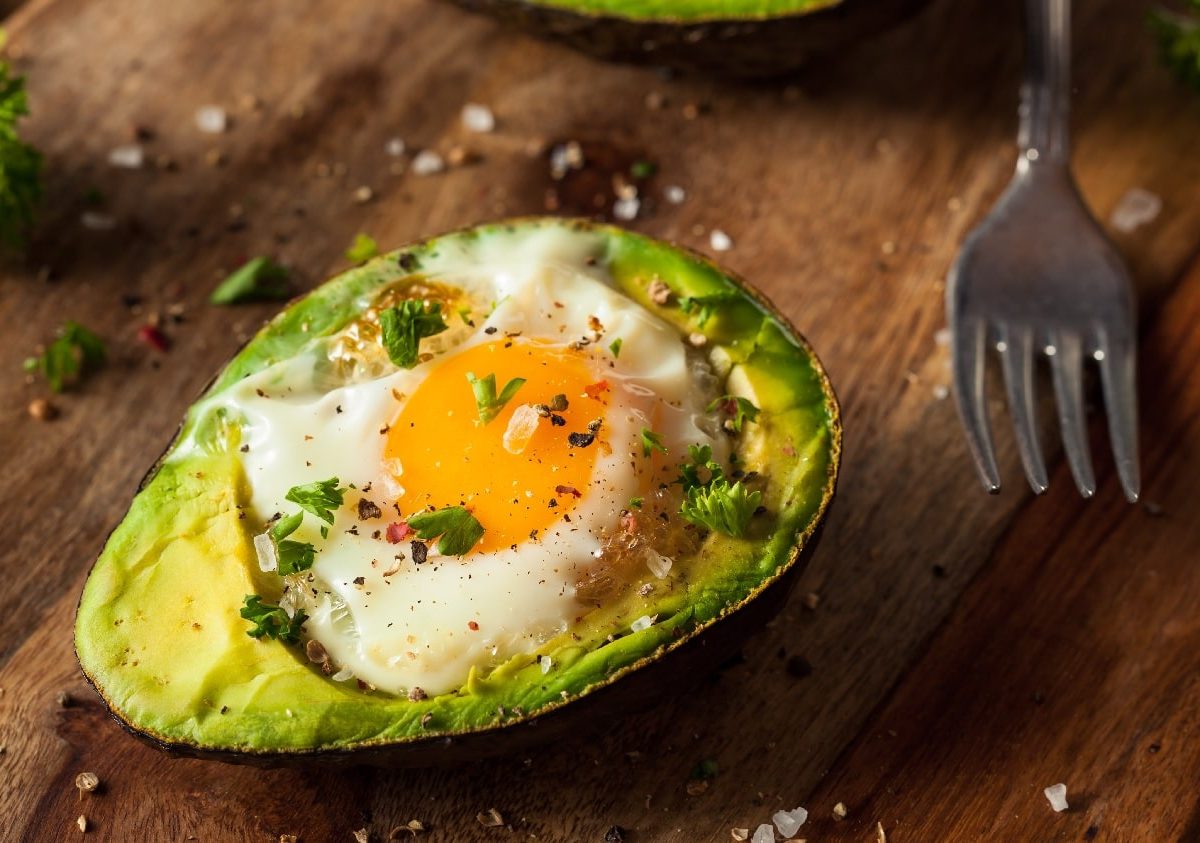 Eine Avocadohälfte mit einem Spiegelei gefüllt auf einem rustikalen Holzbrett. Daneben einiges an Kräutern und Gewürzen.