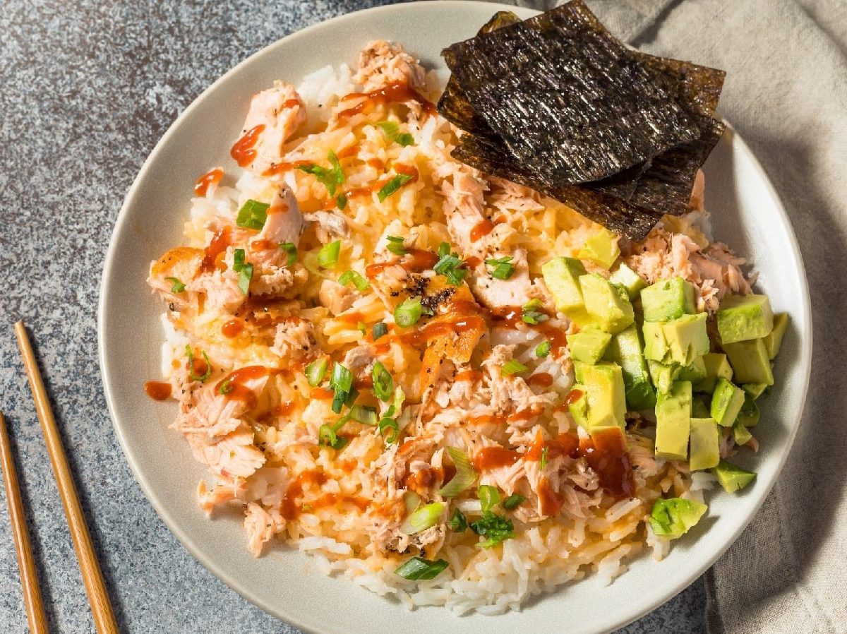 Ein Teller mit Bang Bang Salmon, Avocado und Nori-Blätter auf einem weißen Teller neben einem Paar Stäbchen.