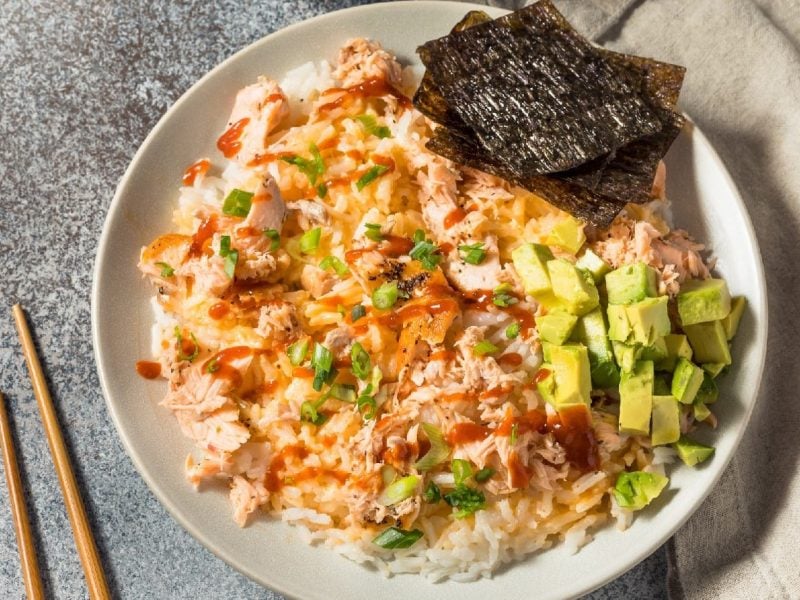 Ein Teller mit Bang Bang Salmon, Avocado und Nori-Blätter auf einem weißen Teller neben einem Paar Stäbchen.