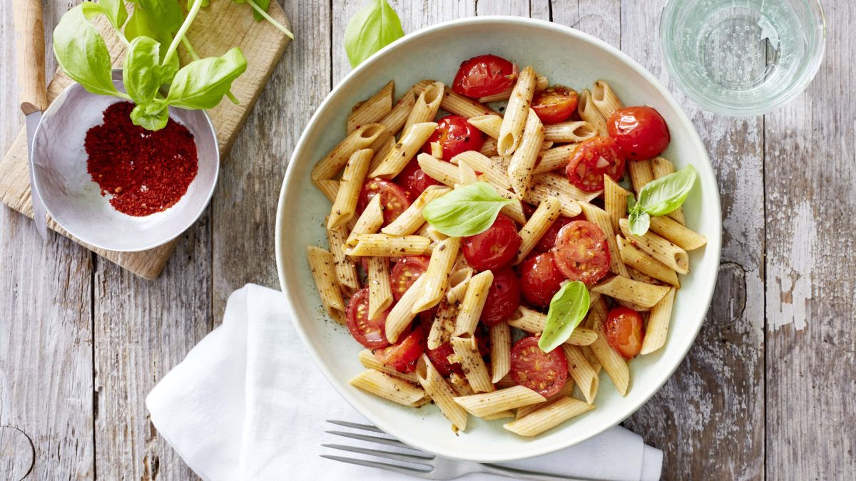 Weißer Teller mit Penne, Tomaten und Basilikum neben einer Schüssel mit Chiliflocken vor hellem Holzhintergrund