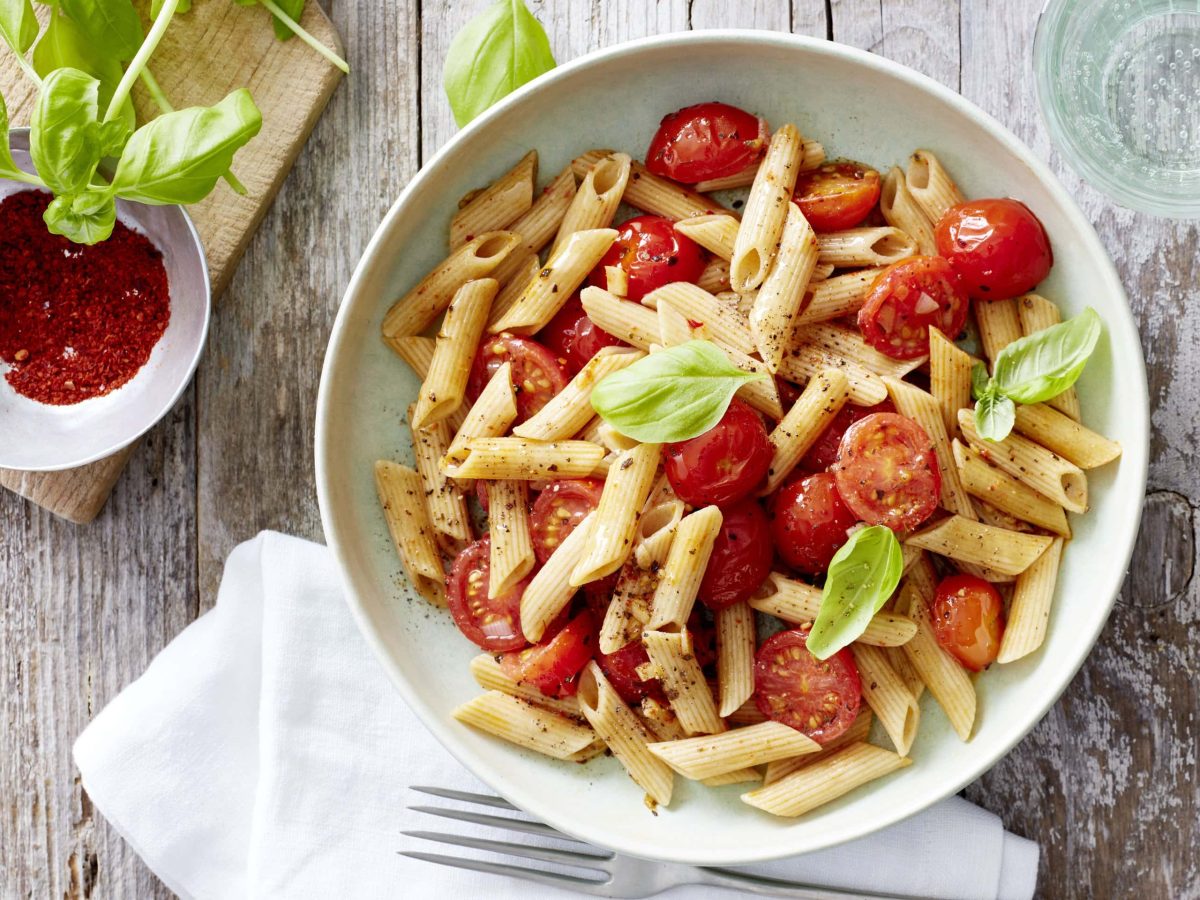 Weißer Teller mit Penne, Tomaten und Basilikum neben einer Schüssel mit Chiliflocken vor hellem Holzhintergrund