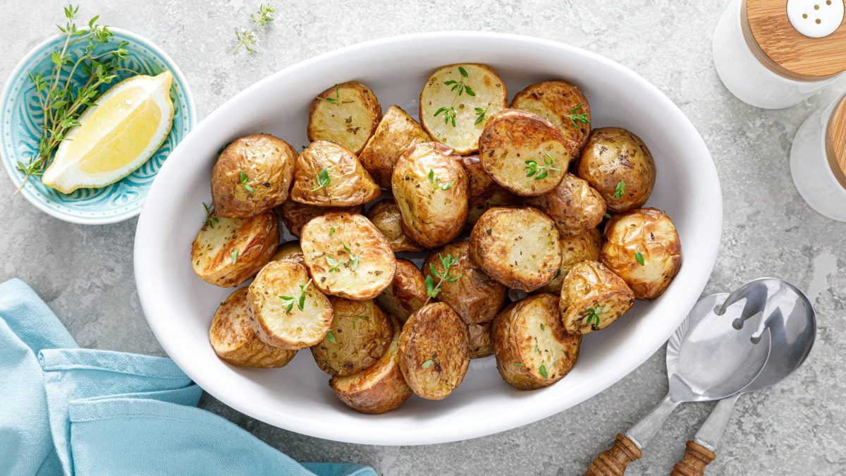 Eine Auflaufform mit Bratkartoffeln im Backofen zubereitet, bestreut mit frischen Kräutern, daneben Salatbesteck, eine Schale mit Zitrone und Kräutern sowie Gewürzstreuer, alles Draufsicht.