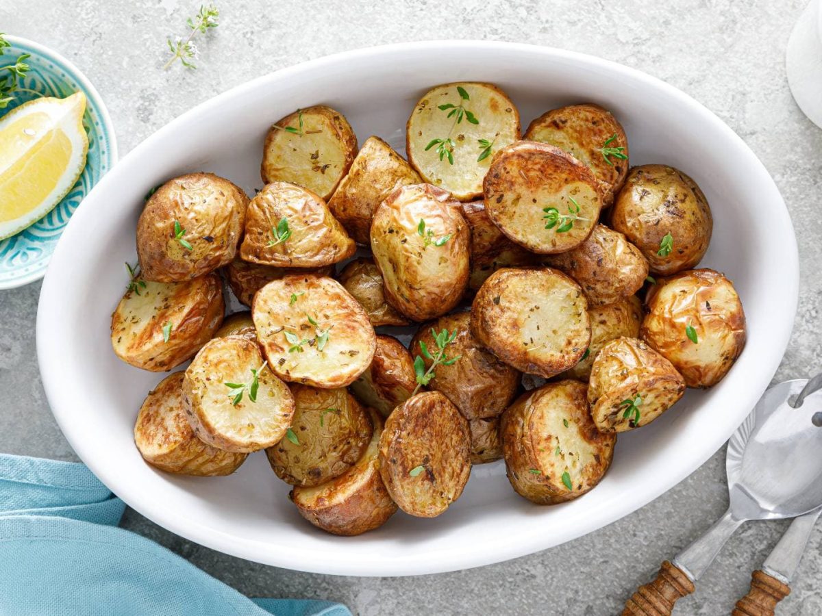 Eine Auflaufform mit Bratkartoffeln im Backofen zubereitet, bestreut mit frischen Kräutern, daneben Salatbesteck, eine Schale mit Zitrone und Kräutern sowie Gewürzstreuer, alles Draufsicht.