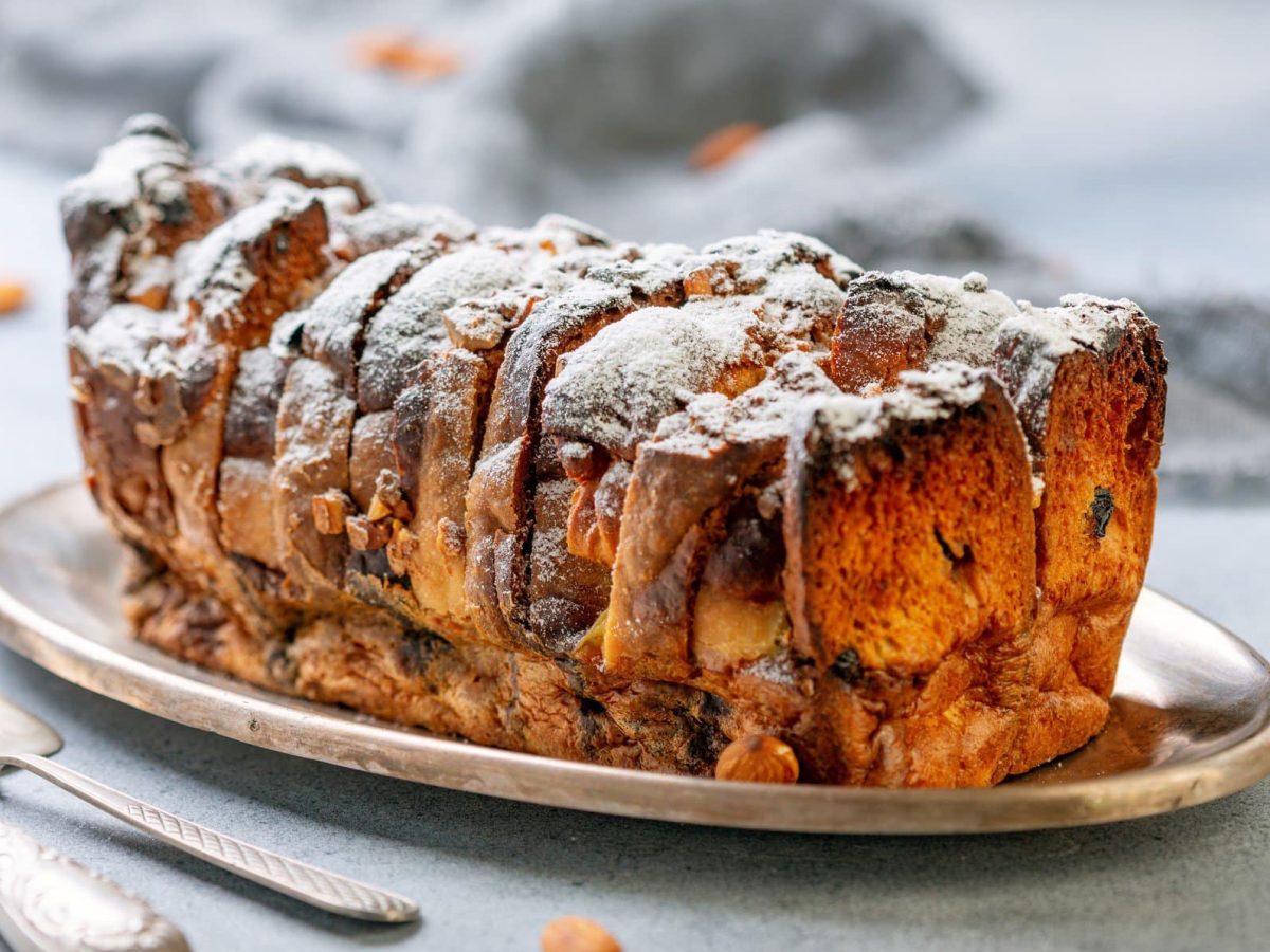 Brotpudding mit Ahornsirup, Banane und Nüssen auf weißem Teller. Im Bild Bestecke und Nüsse. Frontalansicht.