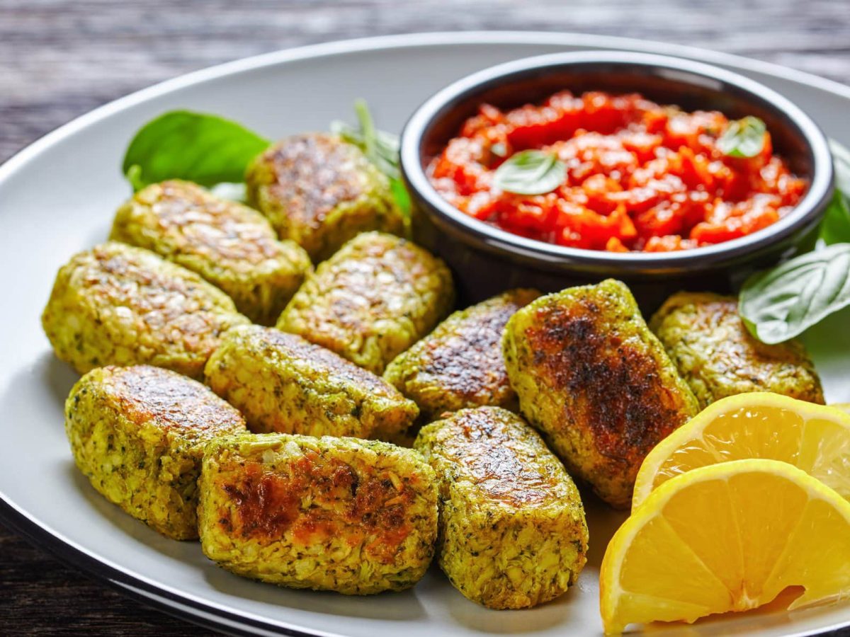 Ein weißer Teller mit den Brokkoli Tots und etwas Basilikum, Zitrone und dem Tomatendip.