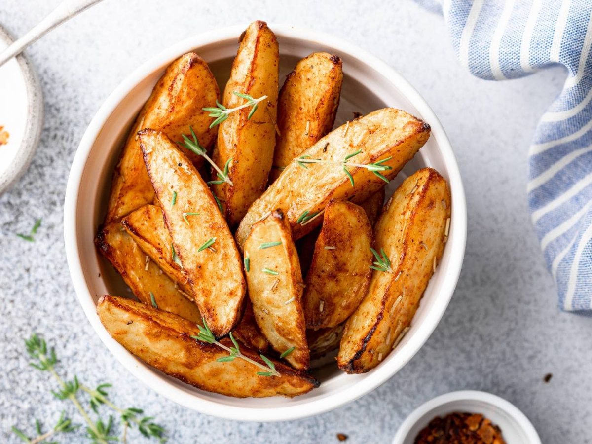 Eine Schale Chilikartoffeln mit Ahornsirup und frischen Kräutern bestreut in der Draufsicht, daneben eine Schale mit Sour Cream, eine mit Chiliflocken und ein Geschirrtuch.