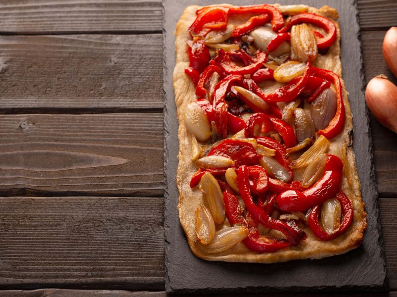 Coca Mallorquina Coca Pizza auf Schieferplatte und Holztisch. Daneben Schalotten. Aufnahme aus der Vogelperspektive.