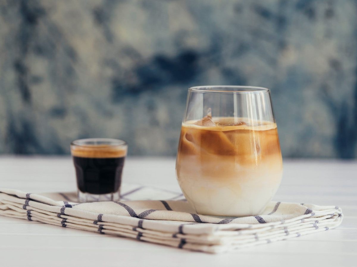 Cold Johnny Eiskaffee in einem Glas mit Espresso auf Tuch und hellem Tisch vor blau-weißem Hintergrund. Frontalansicht.