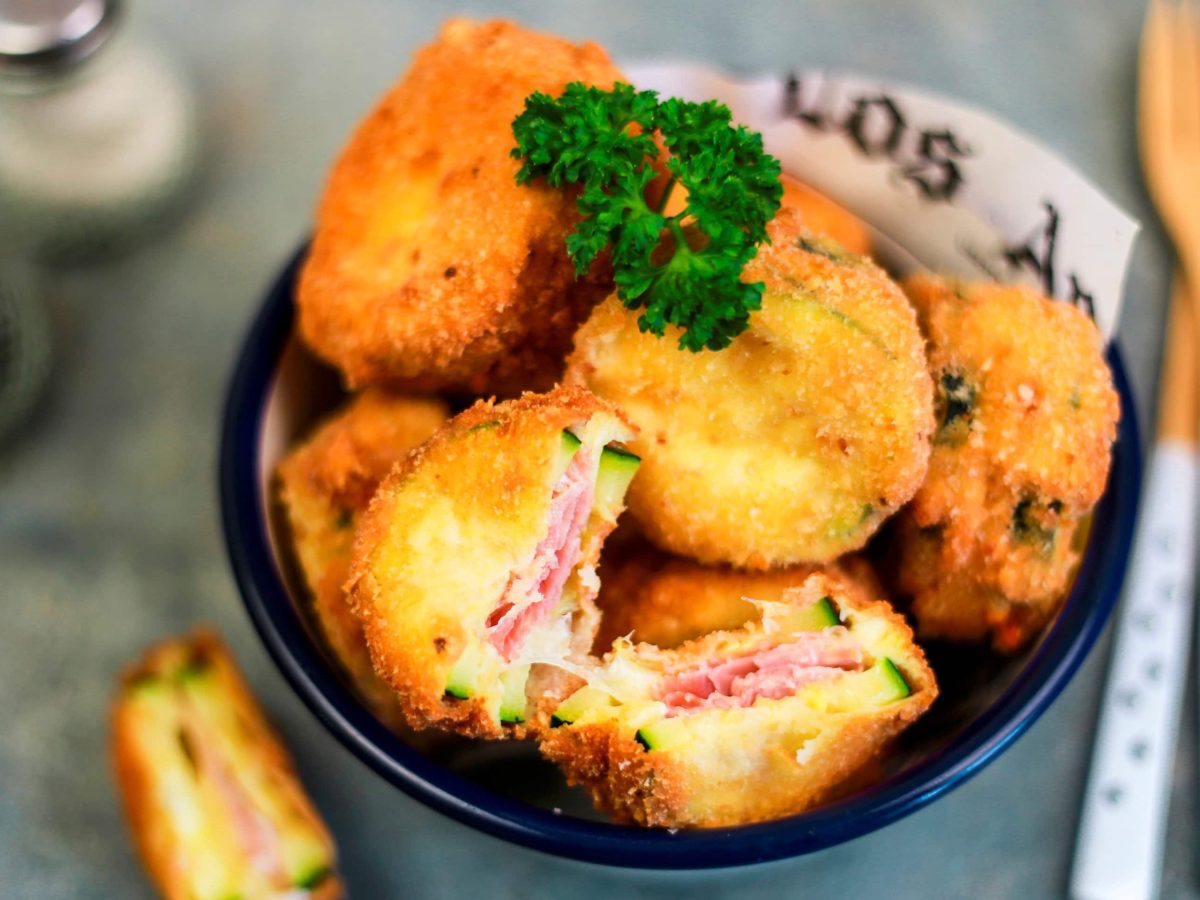 mehrere Cordon Bleu Schnitzel in einer dunklen Schüssel garniert mit Petersilie vor dunklem Hintergrund mit Besteck und Streuern im Hintergrund