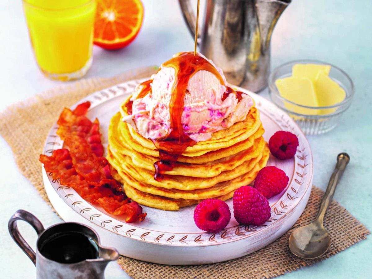 Ein Pancake-Turm mit American Breakfast Eis aus dem Kochbuch "Eis aus dem Wunderland", daneben frische Himbeeren und gebratener Speck, außerdem ein Glas O-Saft, Kännchen und ein Löffel.