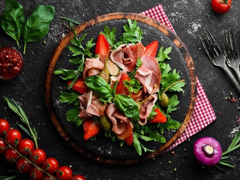 Auf einem Tisch steht ein Erdbeer-Rucola-Salat mit Schinken. Um den Teller liegen Tomaten, Basilikumblätter und Knoblauch.