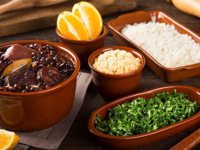 Feijoada in Tonform auf braunem Untergrund. Daneben Beilagen in Tonformen. Holz als Deko im Bild. Frontalansicht.