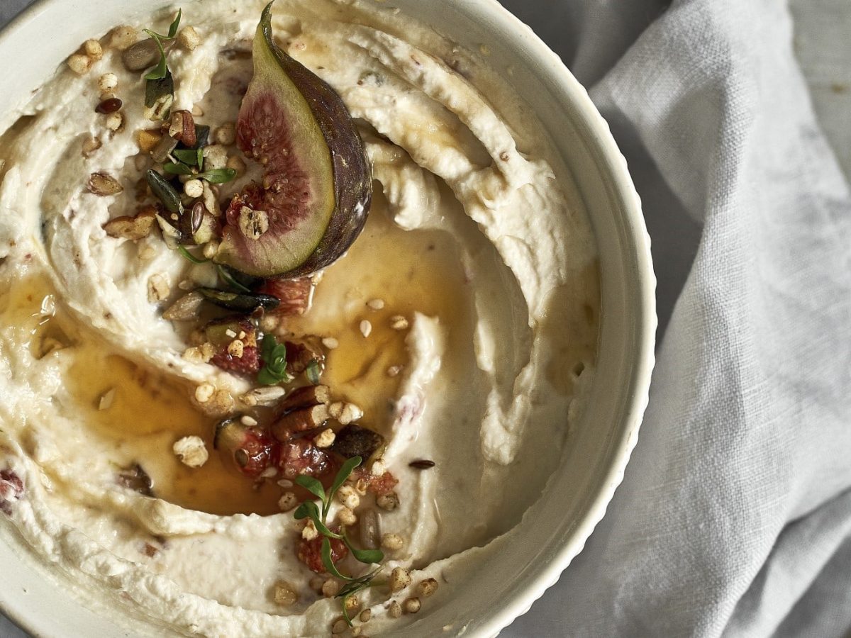 Frischkäsedip mit Feigen und angerösteten Macadamia-Nüssen, übergossen mit Honig und serviert in einer weißen Schale auf hellem Untergrund.