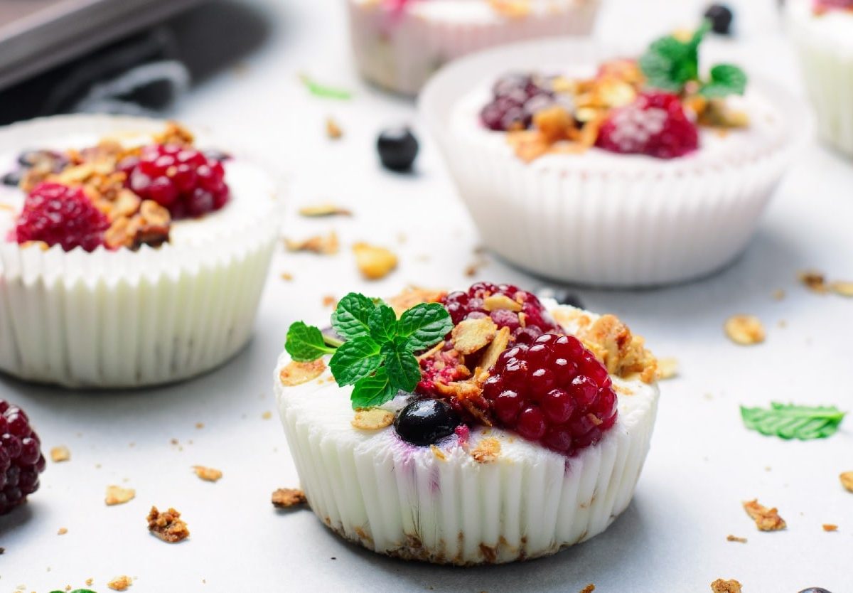 Mehrere Frozen Yoghurt Cups auf einem weißem Tisch. Darauf verteilteinige Haferflocken, Beeren und Minze.